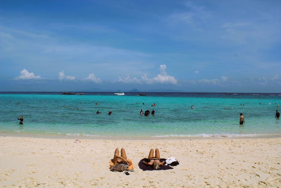 Sunny beach scene in Thailand with people sunbathing and swimming in turquoise waters.