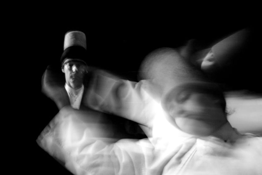 A whirling dervish in mid-spin, arms outstretched—one hand pointed upward to receive divine grace, the other turned downward to channel it into the world.