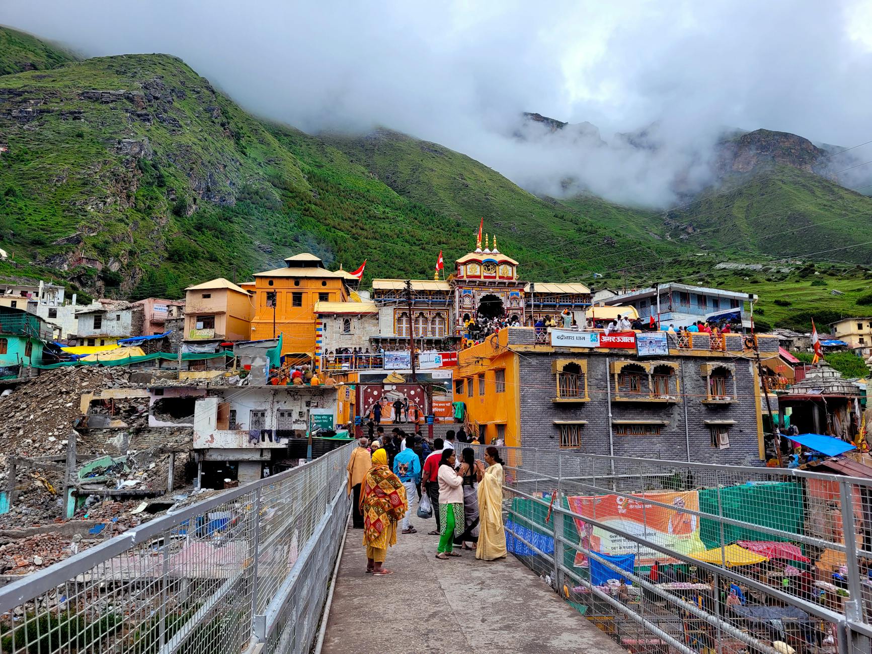 Vibrant view of Badrinath Temple surrounded by lush mountains and visitors crossing a bridge.