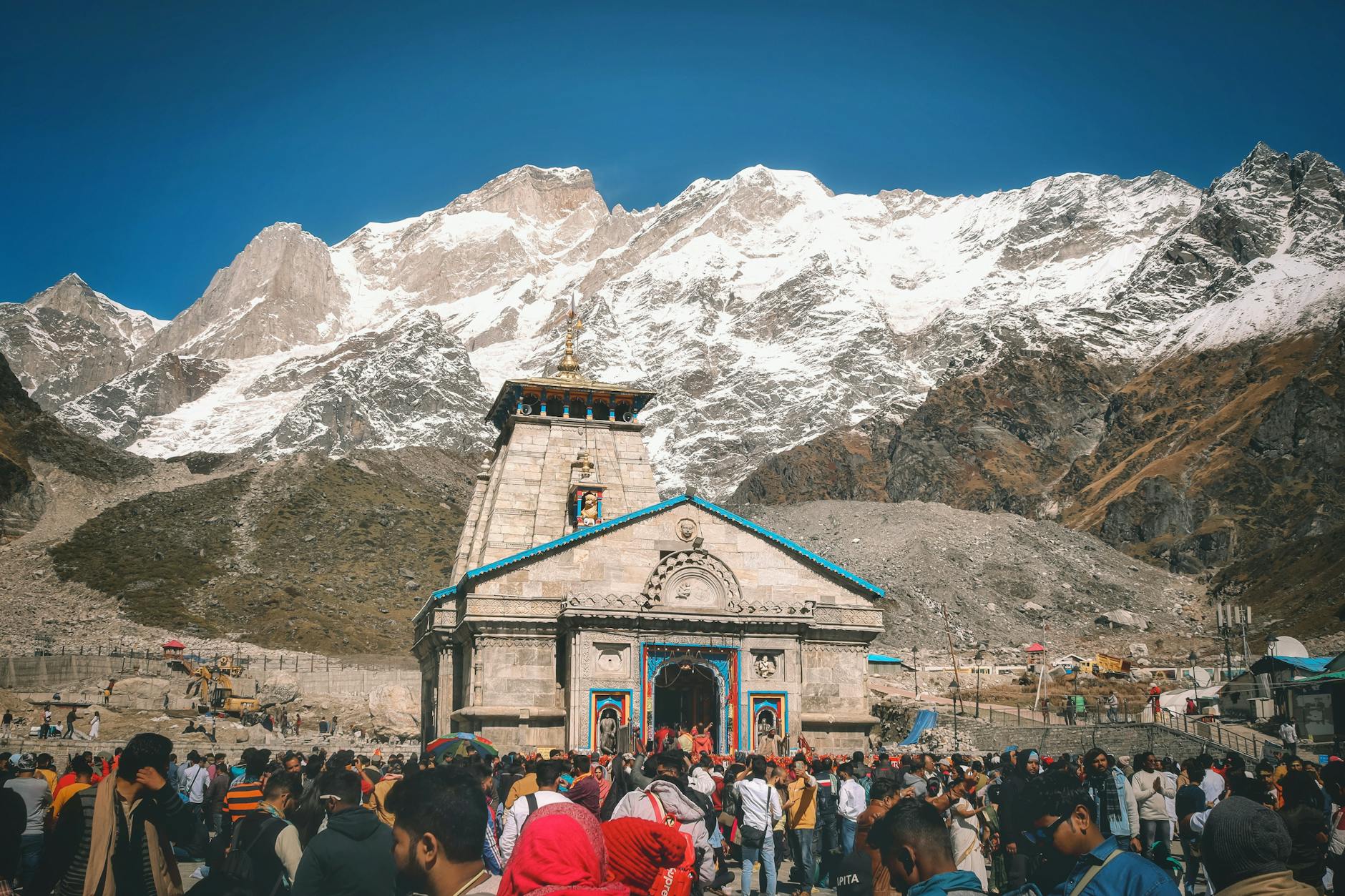 A crowd of pilgrims at Kedarnath Temple with snowcapped Himalayan peaks in the background.