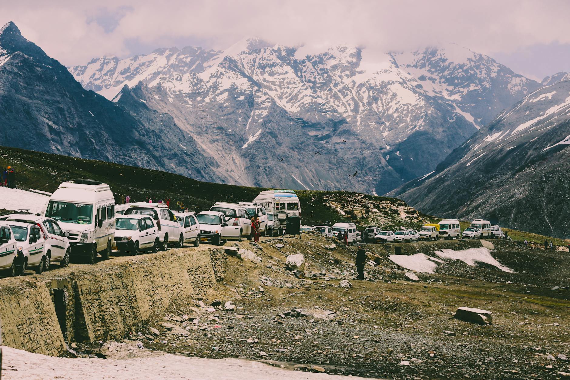 Vehicles lined up on a snowy mountain road, surrounded by majestic peaks and clear sky.