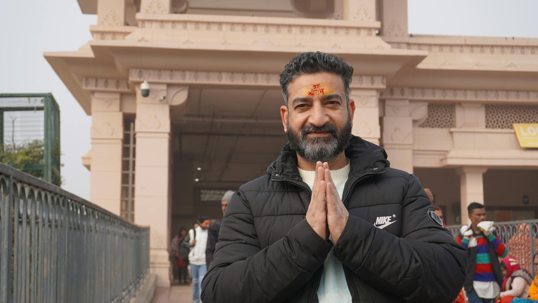 A man dressed warmly performs a Namaste gesture outside a traditional temple.