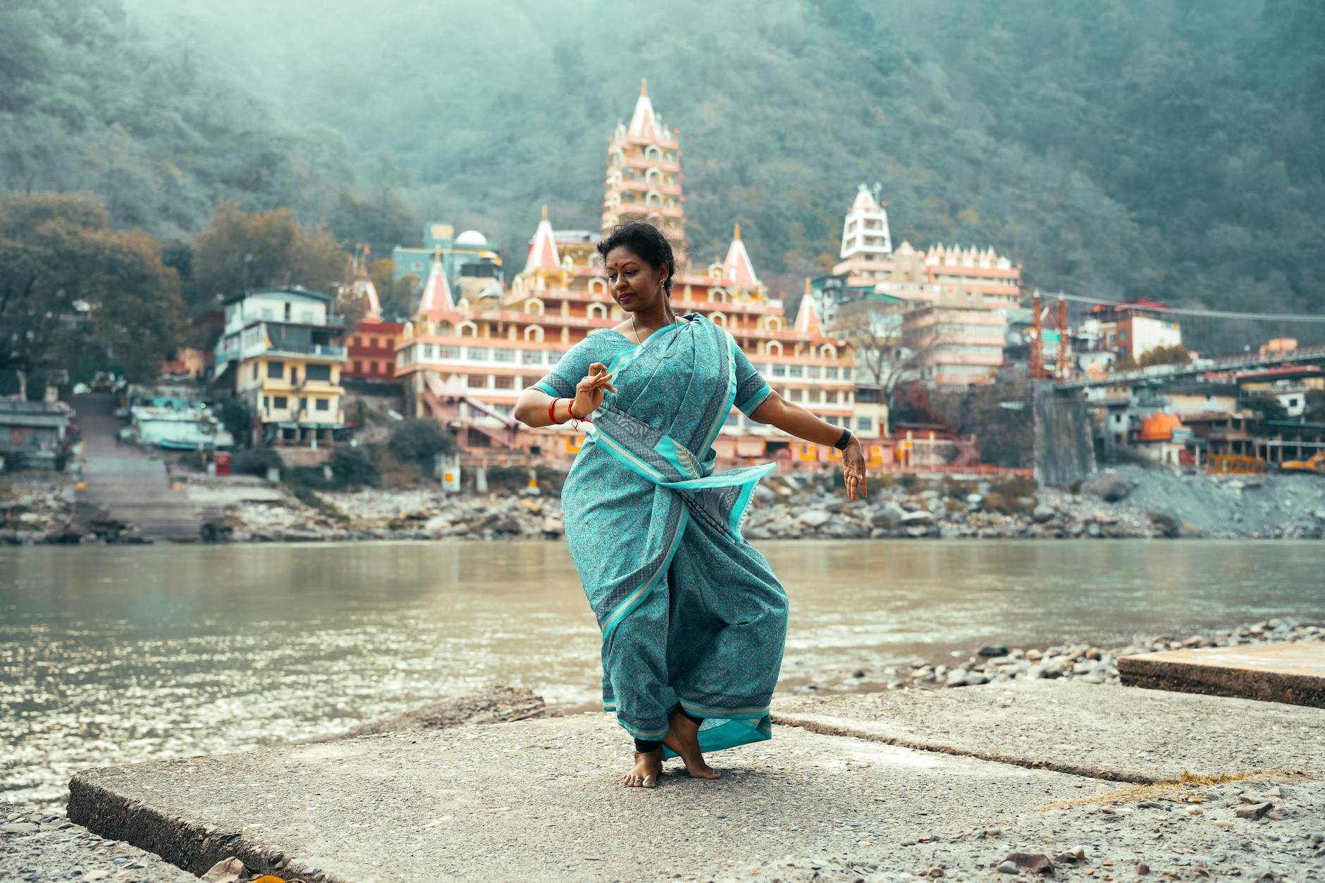 Graceful woman in traditional attire dancing by river in Rishikesh with scenic temples in background.