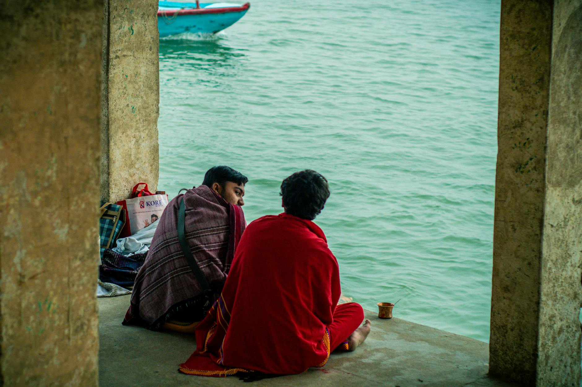 Two men wrapped in shawls sitting by the Ganges River in Varanasi, India, enjoying a peaceful moment.