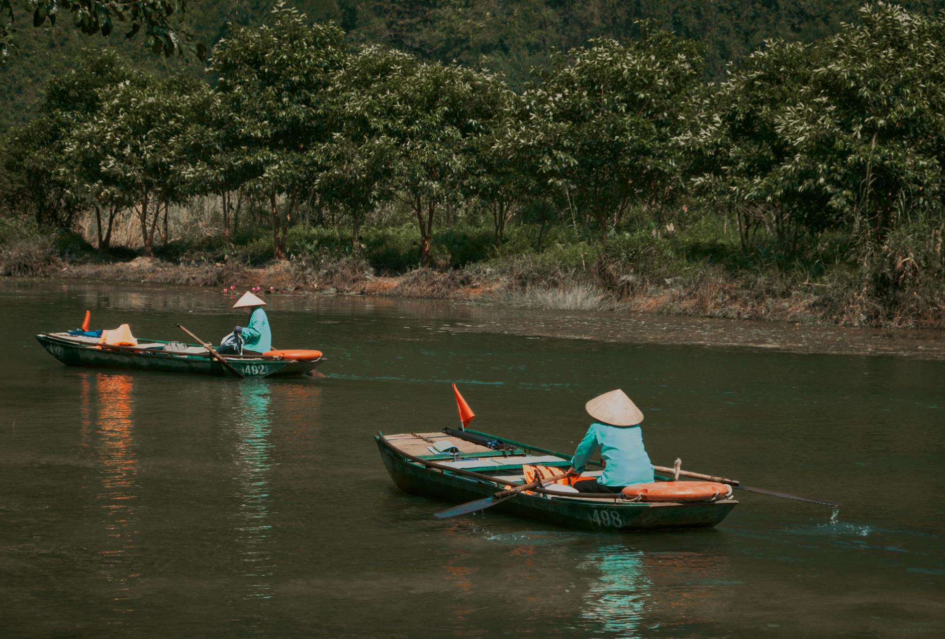 Two people row boats on a tranquil river wearing traditional conical hats.